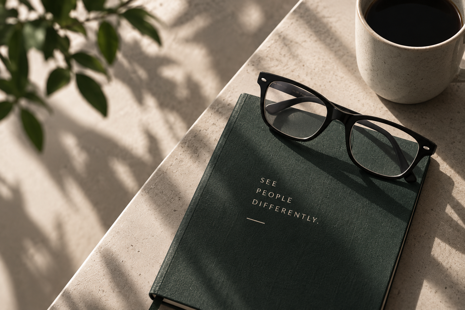 A green notebook reading SEE PEOPLE DIFFERENTLY with glasses resting on it, in raking sunlight